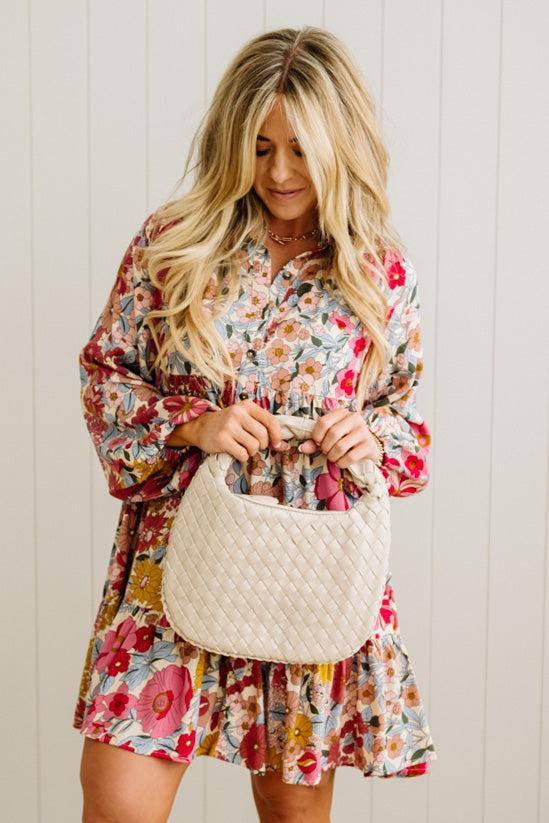 Woman in a floral dress holding a beige handbag against a white wall.