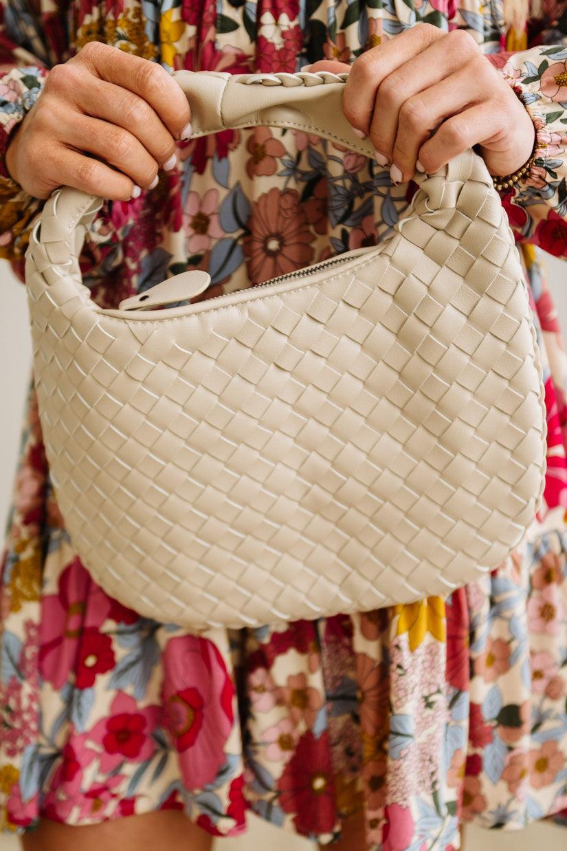 Beige woven handbag held by a person wearing a floral dress.