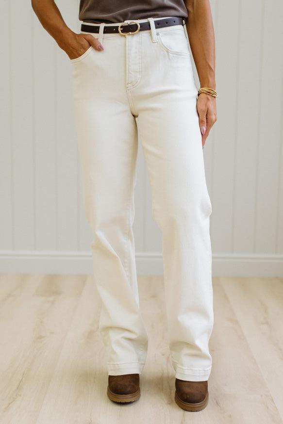 A person wearing off-white straight-leg jeans with a black belt, paired with brown suede boots, standing in a room with light wood floors and white paneled walls.