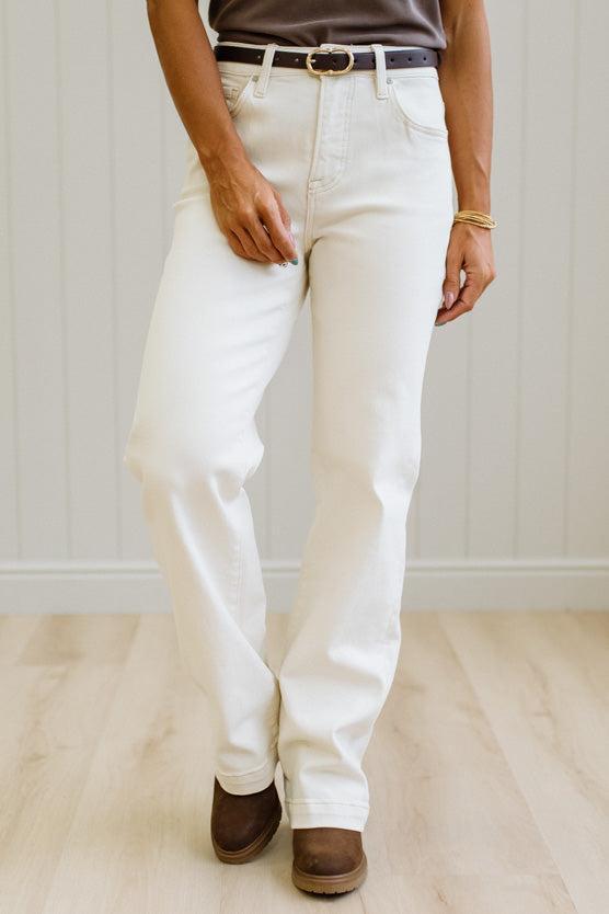 A person wearing off-white straight-leg jeans with a dark belt, paired with brown boots, standing on a light wood floor against a white paneled wall.