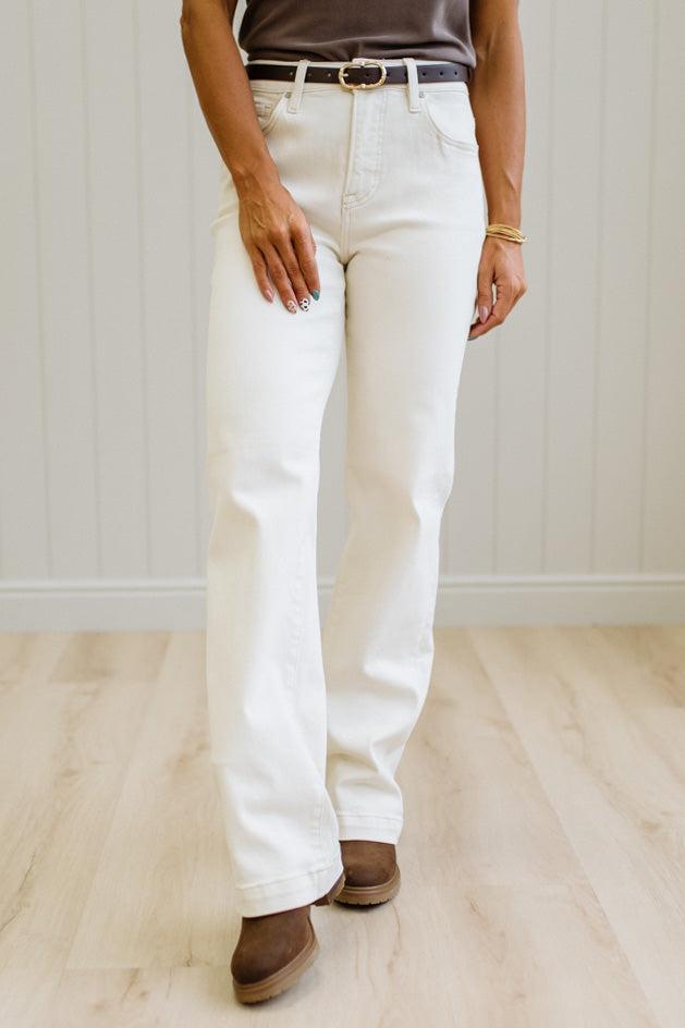 A person wearing off-white straight-leg jeans with a brown belt and brown suede boots, standing on a light wood floor against a white paneled wall.