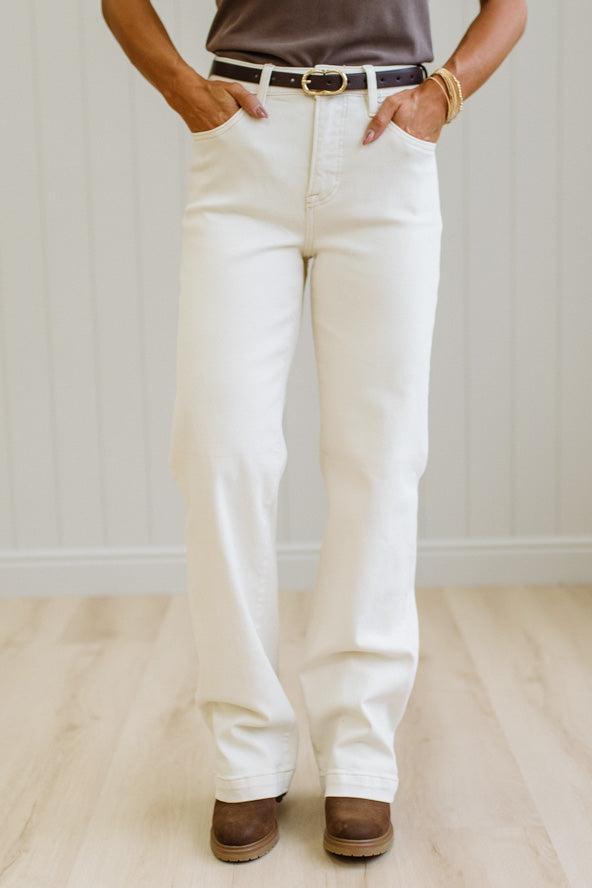 A person wearing off-white straight-leg jeans with a brown belt, paired with a brown top and brown shoes, standing in a room with light wood floors and white paneled walls.