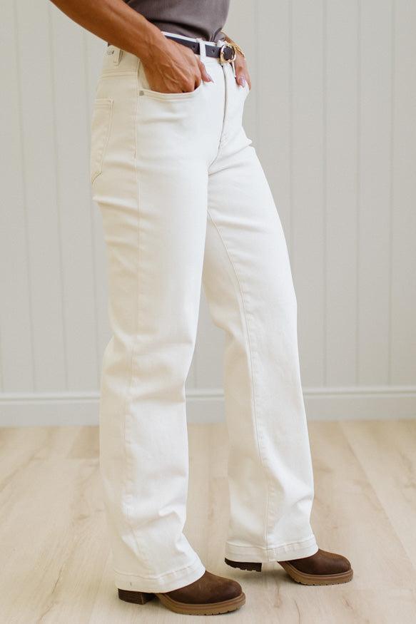 A person wearing off-white or cream wide-leg jeans with brown shoes, standing in a room with light wood flooring and a white paneled wall.