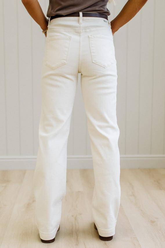 A person shown from the back wearing off-white or cream-colored straight-leg jeans, paired with a dark brown top and dark brown shoes, standing on a light wood floor against a white paneled wall.