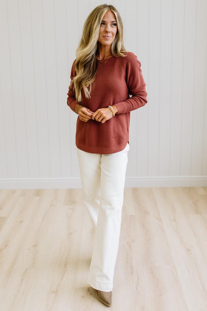 A woman modeling a rust-colored long-sleeve knit top and off-white wide-leg pants in a bright room with light wood floors and white paneled walls.