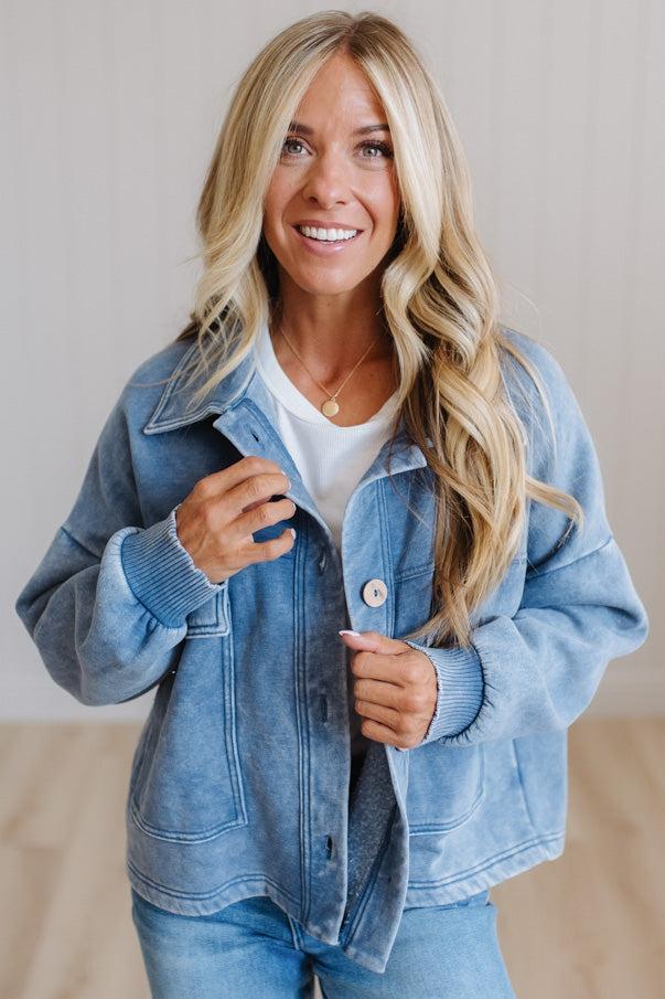 A smiling blonde woman wearing a light blue washed denim jacket over a white t-shirt and blue jeans, standing against a neutral light beige wall.