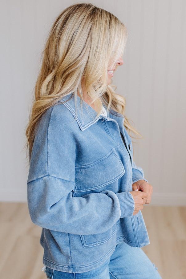 A woman wearing a light blue washed denim jacket and matching blue jeans, shown from a side profile in a bright indoor setting with a light-colored wall and wood floor.
