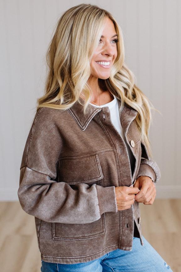 A smiling blonde woman wearing a brown washed button-front jacket over a white top and blue jeans, photographed in a bright indoor setting.
