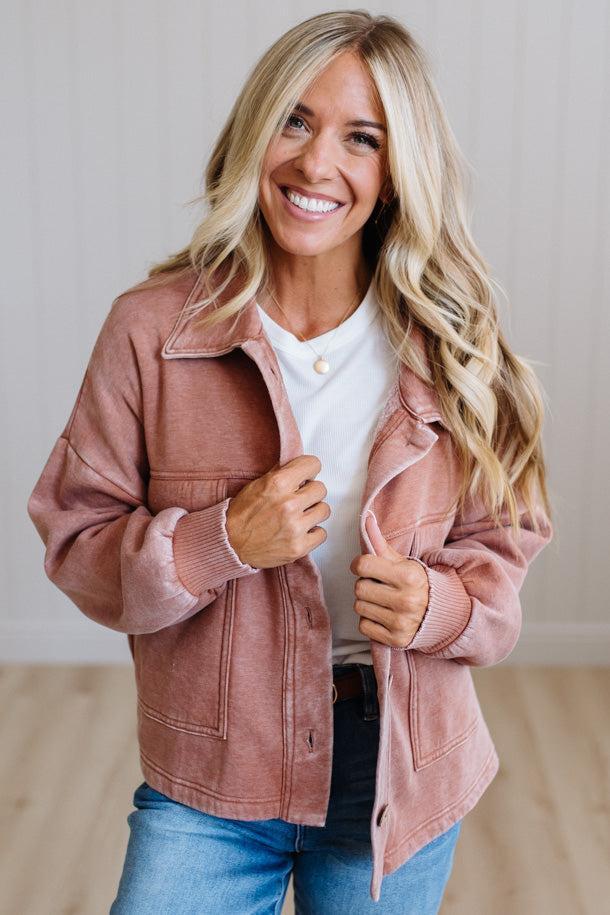 A smiling blonde woman wearing a dusty rose pink corduroy jacket over a white t-shirt and blue jeans, standing in a brightly lit indoor setting.