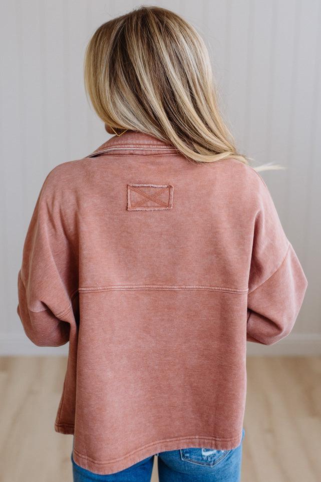 A woman viewed from behind wearing a dusty rose long-sleeve shirt jacket with a small rectangular patch on the upper back, paired with blue jeans.