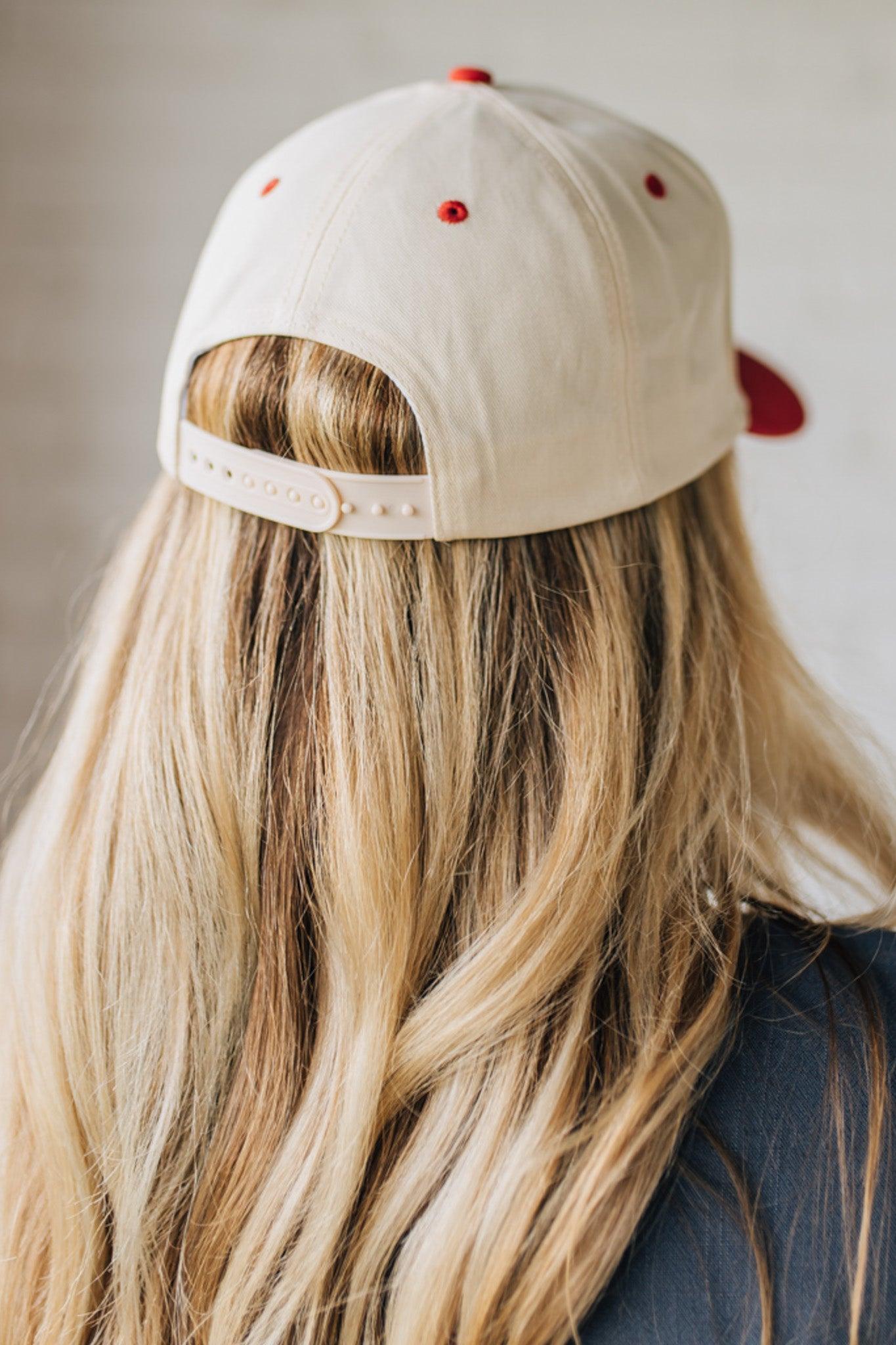 Blonde woman wearing a trucker hat in cream with red brim, and red and blue embroidery at the front "Out of Your League"