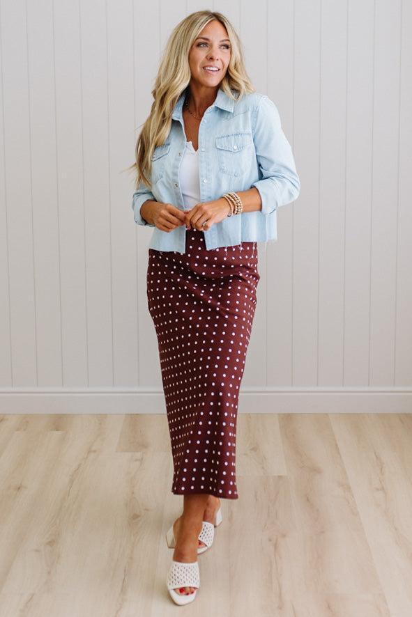Woman wearing a light blue denim jacket over a white shirt and burgundy polka dot skirt on a plain background