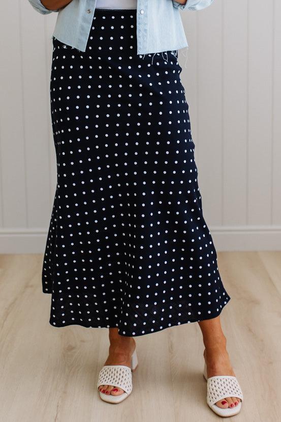 Person wearing a black polka dot skirt with a light blue shirt and white sandals on a wooden floor.