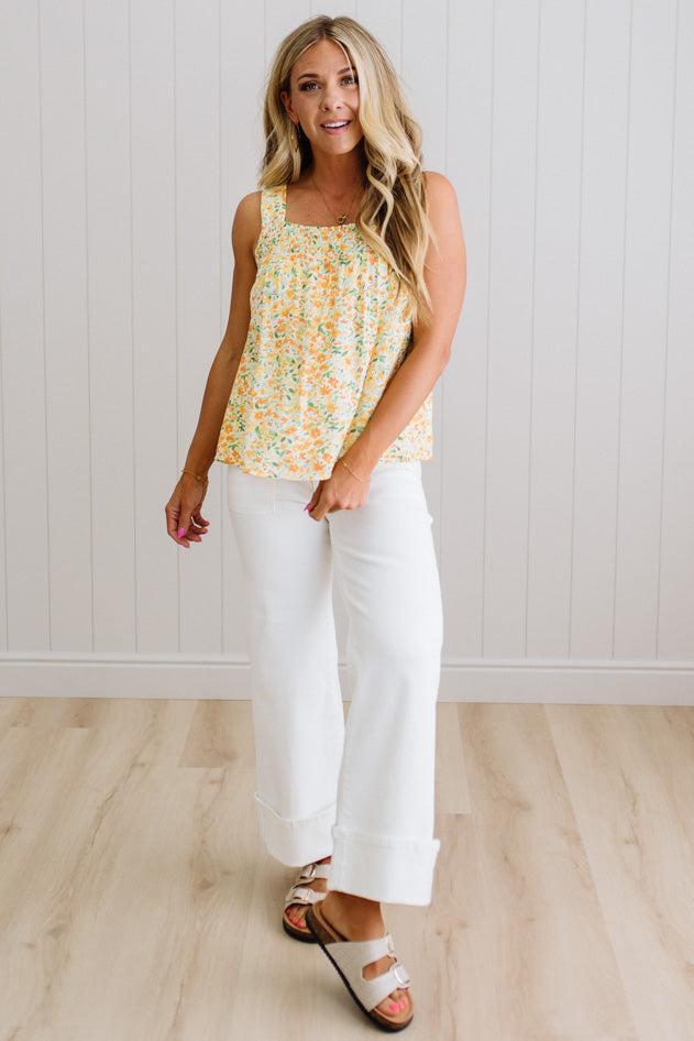 Woman wearing a floral sleeveless top and white pants against a white wall.
