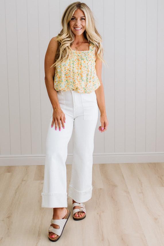 Woman wearing a floral top and white pants standing against a white paneled wall.