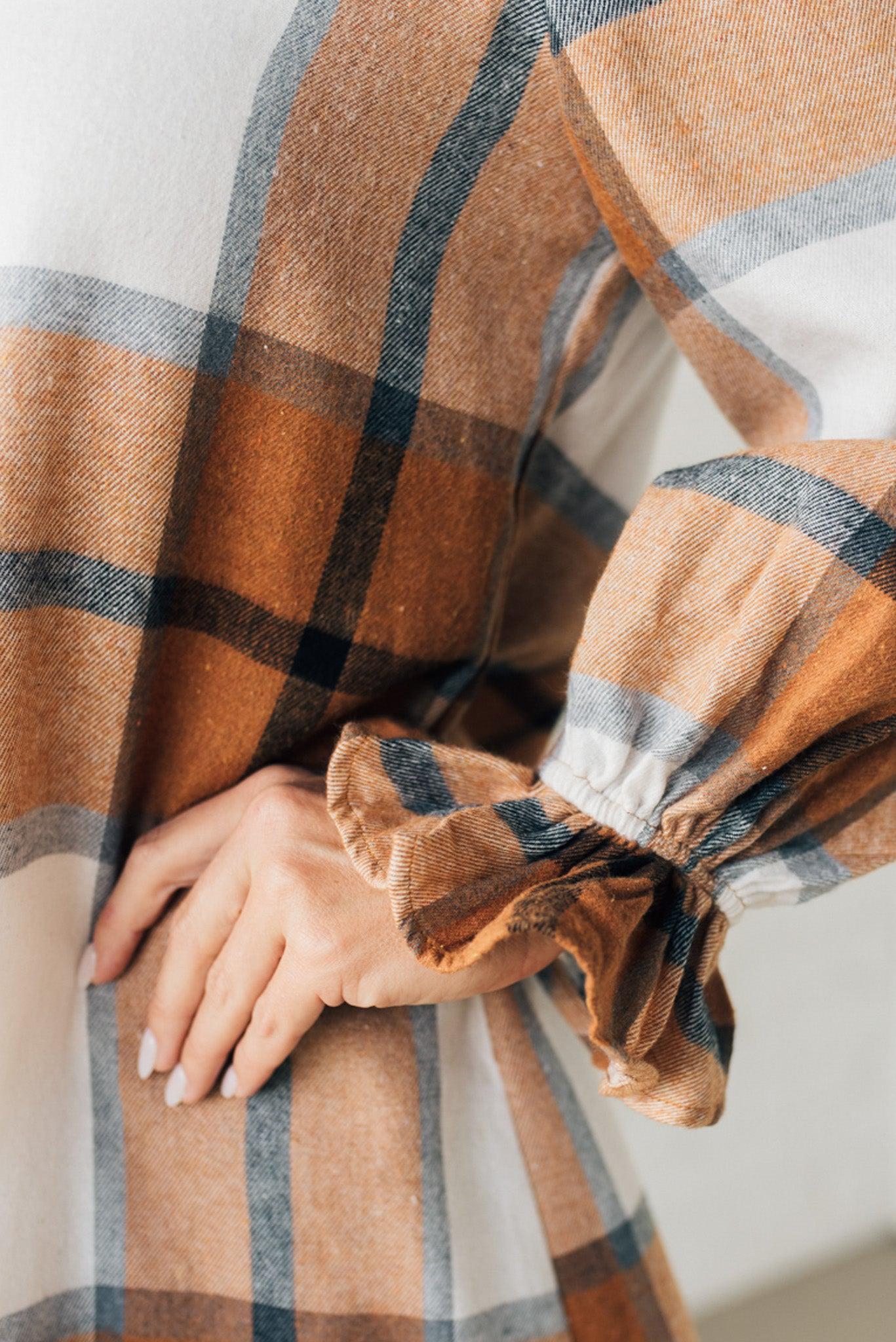 Blonde woman in a fedora wearing a large plaid printed knee length dress with collar, button front, long sleeves with ruffled wrist.
