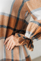 Blonde woman in a fedora wearing a large plaid printed knee length dress with collar, button front, long sleeves with ruffled wrist.