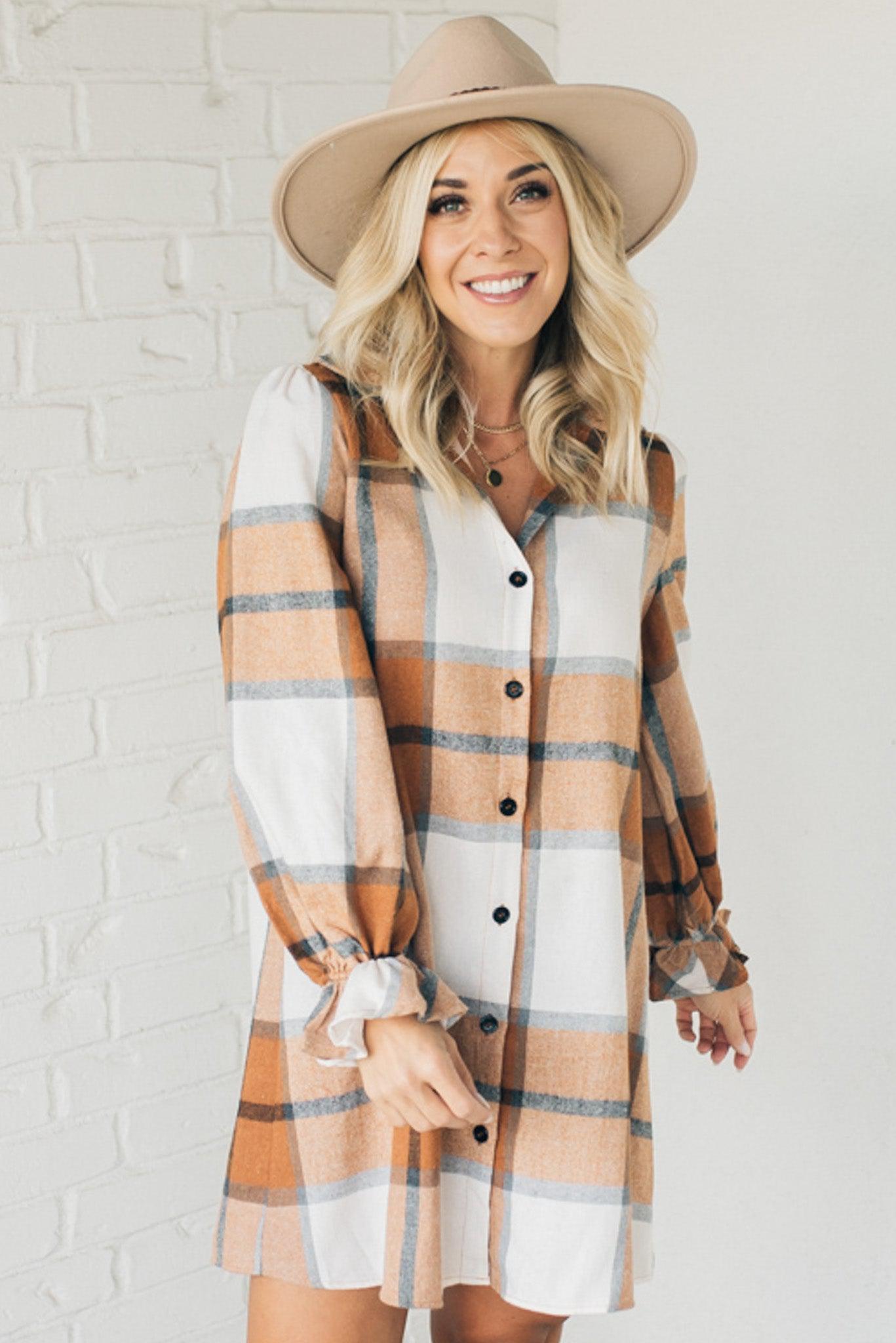 Blonde woman in a fedora wearing a large plaid printed knee length dress with collar, button front, long sleeves with ruffled wrist.