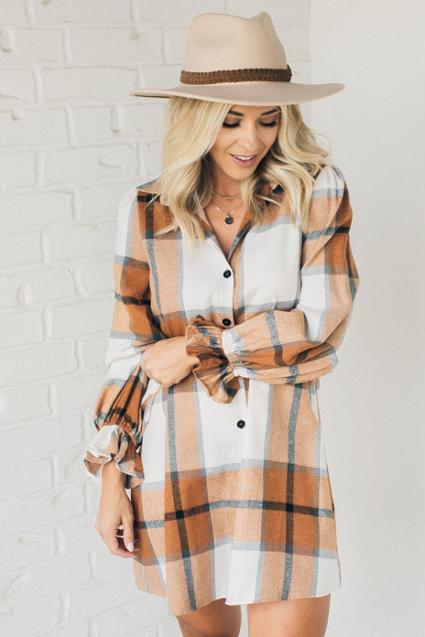 Blonde woman in a fedora wearing a large plaid printed knee length dress with collar, button front, long sleeves with ruffled wrist.