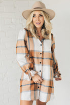 Blonde woman in a fedora wearing a large plaid printed knee length dress with collar, button front, long sleeves with ruffled wrist.