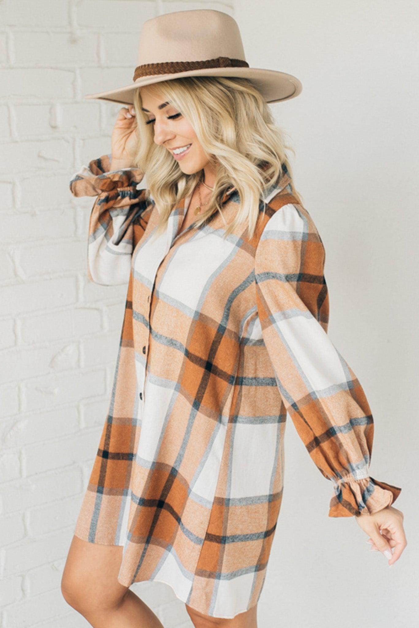 Blonde woman in a fedora wearing a large plaid printed knee length dress with collar, button front, long sleeves with ruffled wrist.