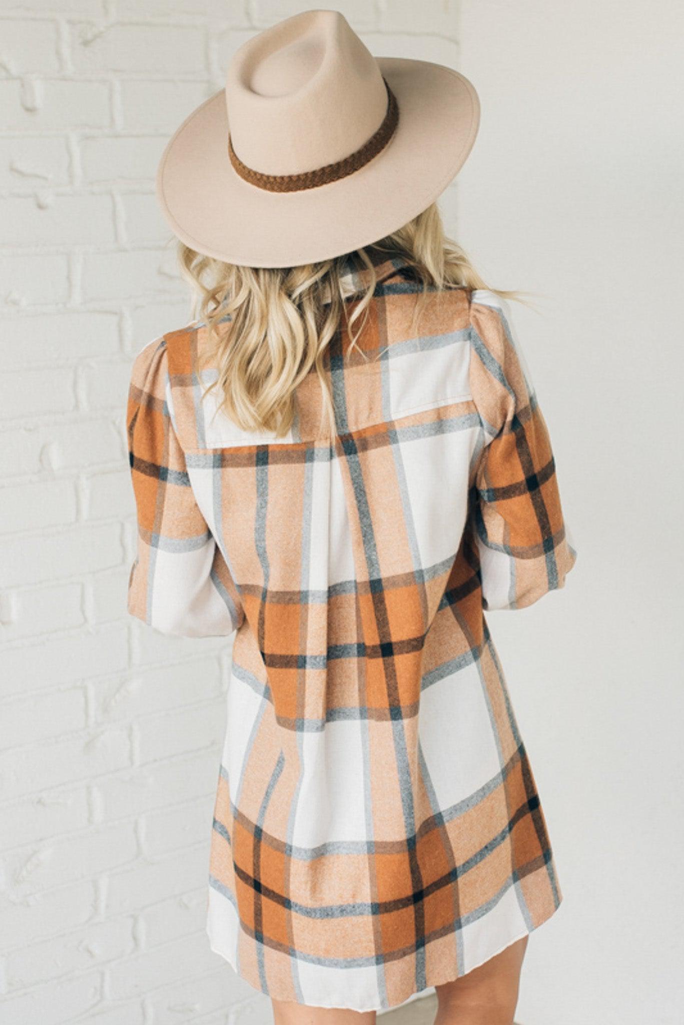 Blonde woman in a fedora wearing a large plaid printed knee length dress with collar, button front, long sleeves with ruffled wrist.