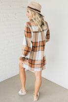 Blonde woman in a fedora wearing a large plaid printed knee length dress with collar, button front, long sleeves with ruffled wrist.