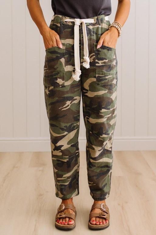 A person wearing camouflage jogger pants with a white drawstring waist, standing in a room with light wood flooring and white paneled walls.