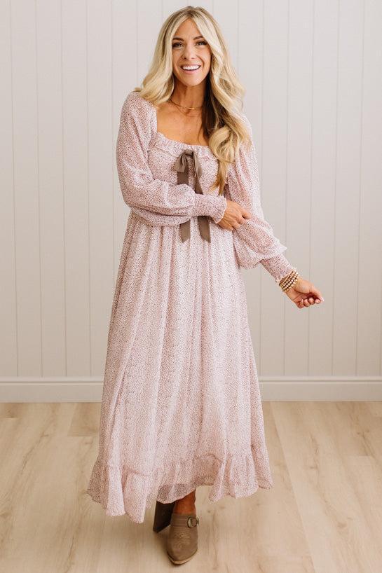 A woman modeling a long-sleeve, square-neck maxi dress in a dusty pink color with a subtle floral pattern and a brown bow at the neckline, standing in front of a white paneled wall and light wood floor.