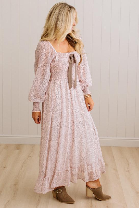 A woman modeling a long-sleeve dusty pink maxi dress with a tie-neck detail, standing in front of a white paneled wall and light wood floor.