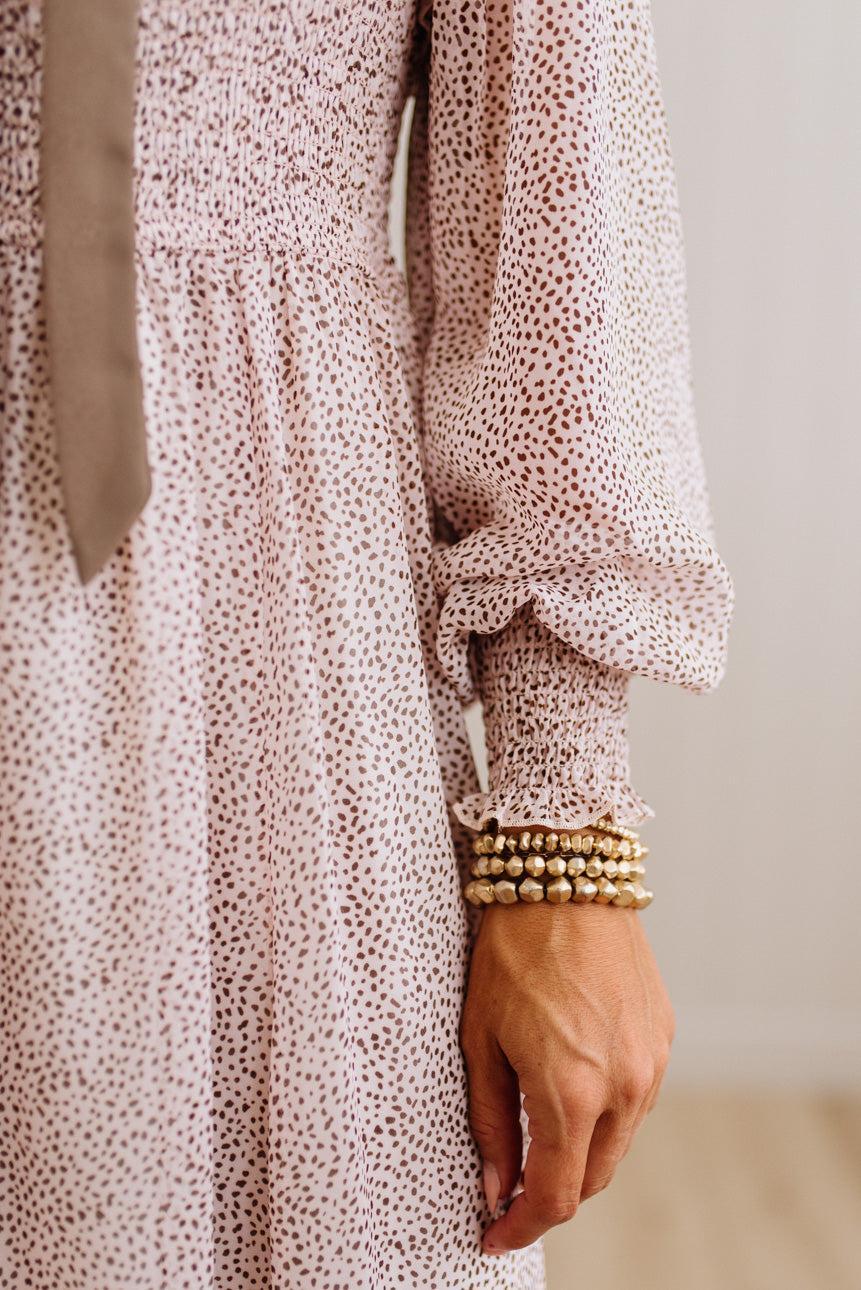 Close-up of a person wearing a light pink long-sleeve dress with a small dark floral print and a tie at the neckline, accessorized with stacked gold beaded bracelets.