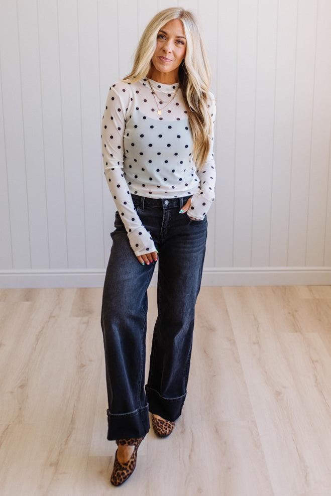Woman wearing a polka dot sweater and jeans standing in a room with light wood flooring and white walls.