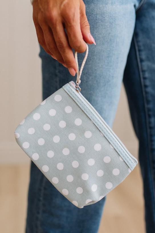 A person holding a small zip pouch with a wrist strap. The pouch is light blue with white polka dots and has a silver zipper.