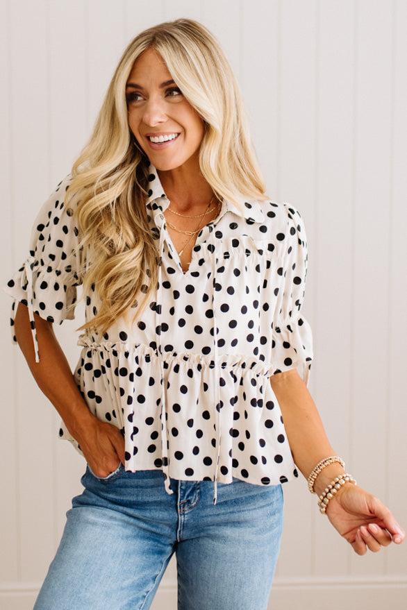 A woman wearing a white short-sleeve blouse with black polka dots and blue jeans, standing against a light beige paneled wall.