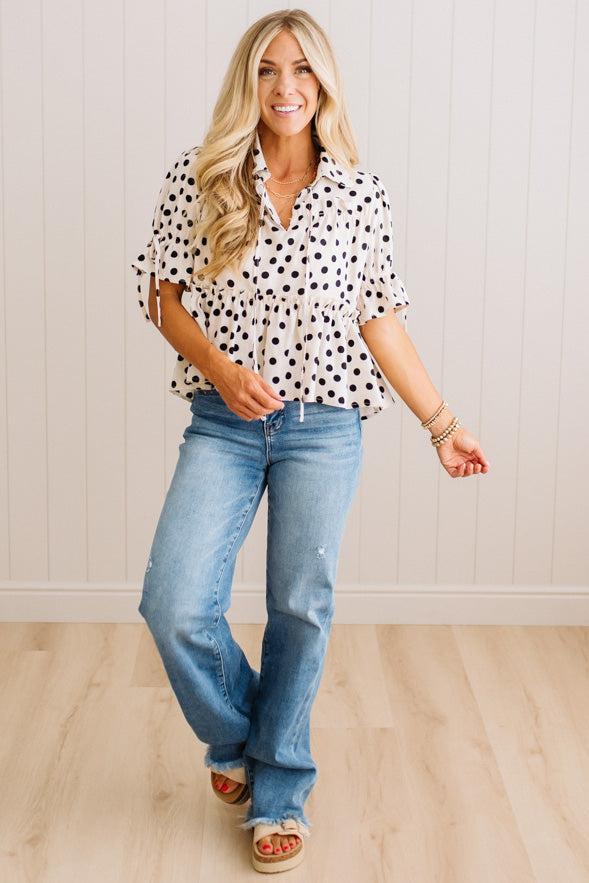 A woman modeling a white short-sleeve blouse with black polka dots, paired with light-wash blue jeans and tan sandals, standing against a white paneled wall and light wood floor.