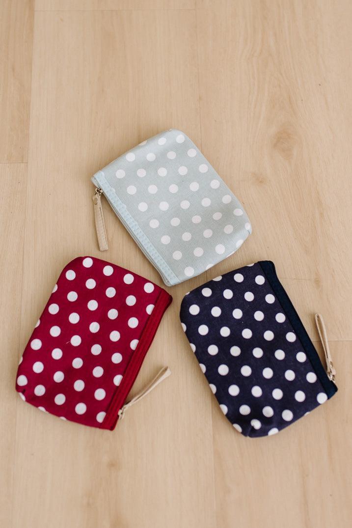 Three small zip pouches with white polka dots are arranged on a light wood floor. One pouch is light blue, one is dark red, and one is dark navy, each with a small gold zipper pull.