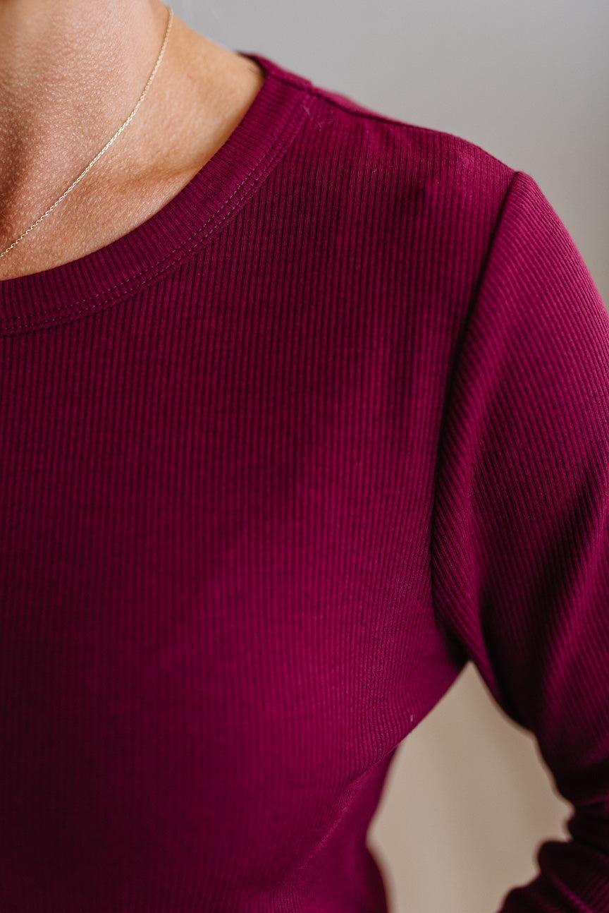 Close-up of a person wearing a ribbed long-sleeve crewneck top in deep burgundy, with a thin gold chain necklace visible against the neck.