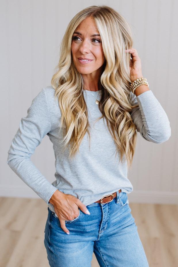 A woman with long blonde wavy hair wearing a light gray long-sleeve henley shirt and blue jeans, standing in a brightly lit indoor room with light wood flooring and white paneled walls.