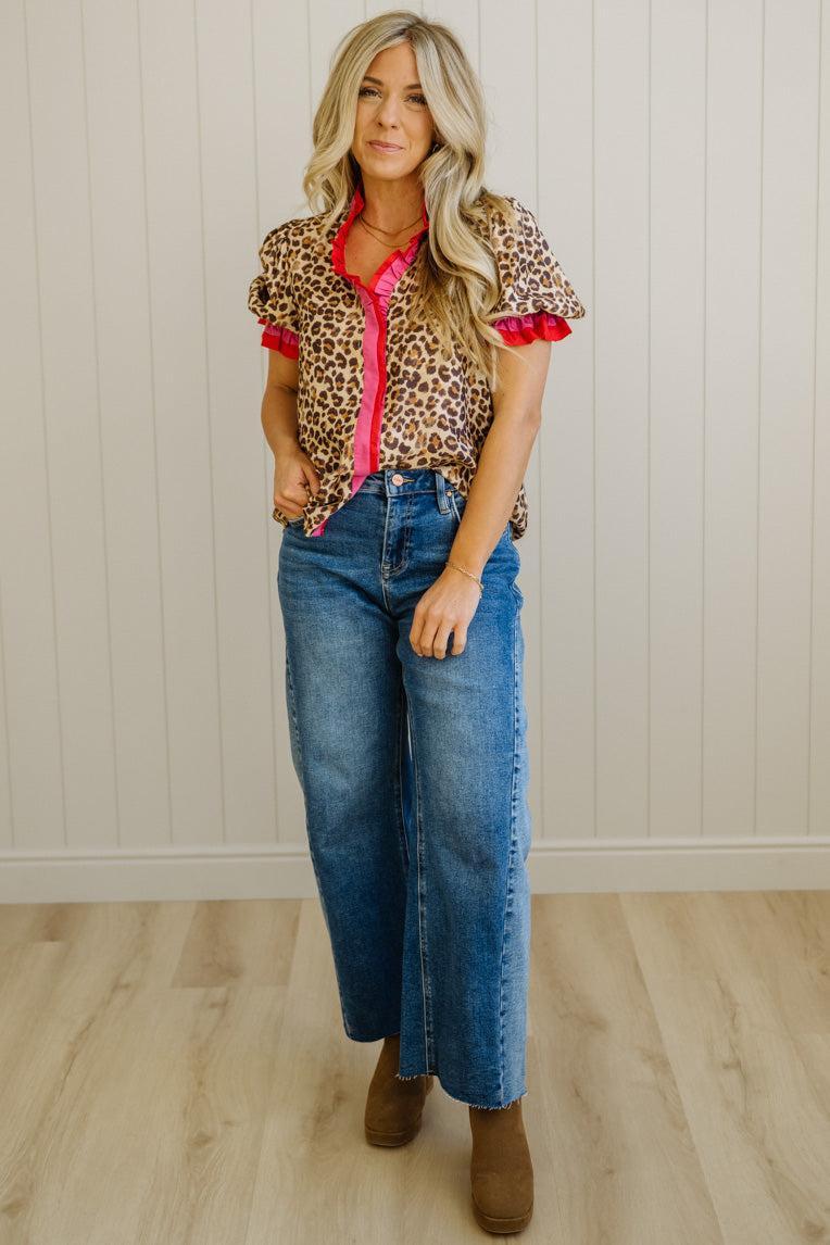 Woman wearing a leopard print shirt with red accents and blue jeans in a room with wooden flooring and white walls.