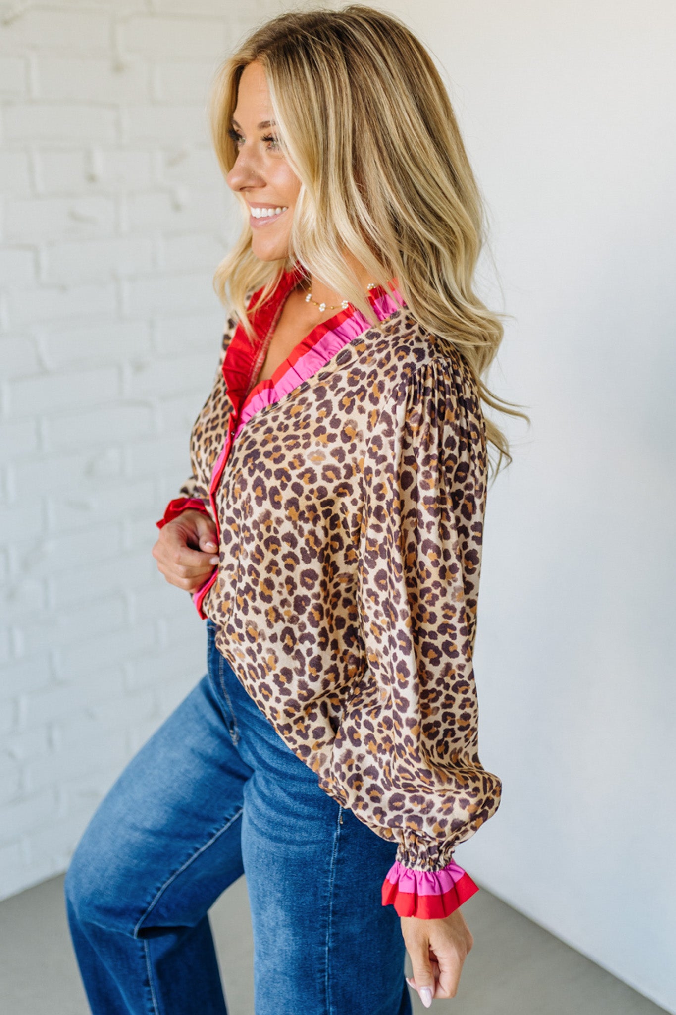 Woman wearing a leopard print blouse with pink trim and blue jeans against a white brick wall.