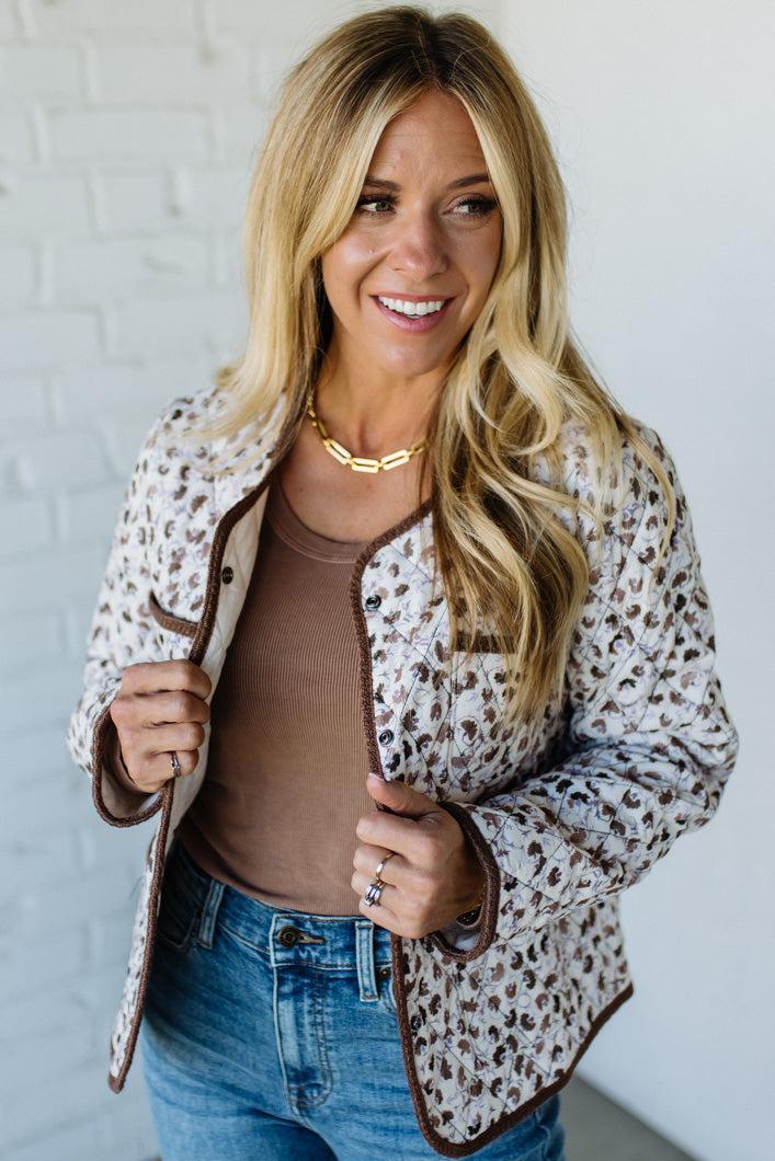 Woman wearing a leopard print jacket over a brown top and blue jeans against a white brick wall.