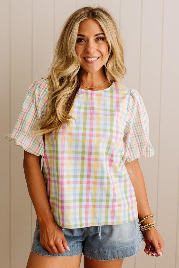 Woman wearing a colorful plaid shirt and denim shorts against a beige wall.