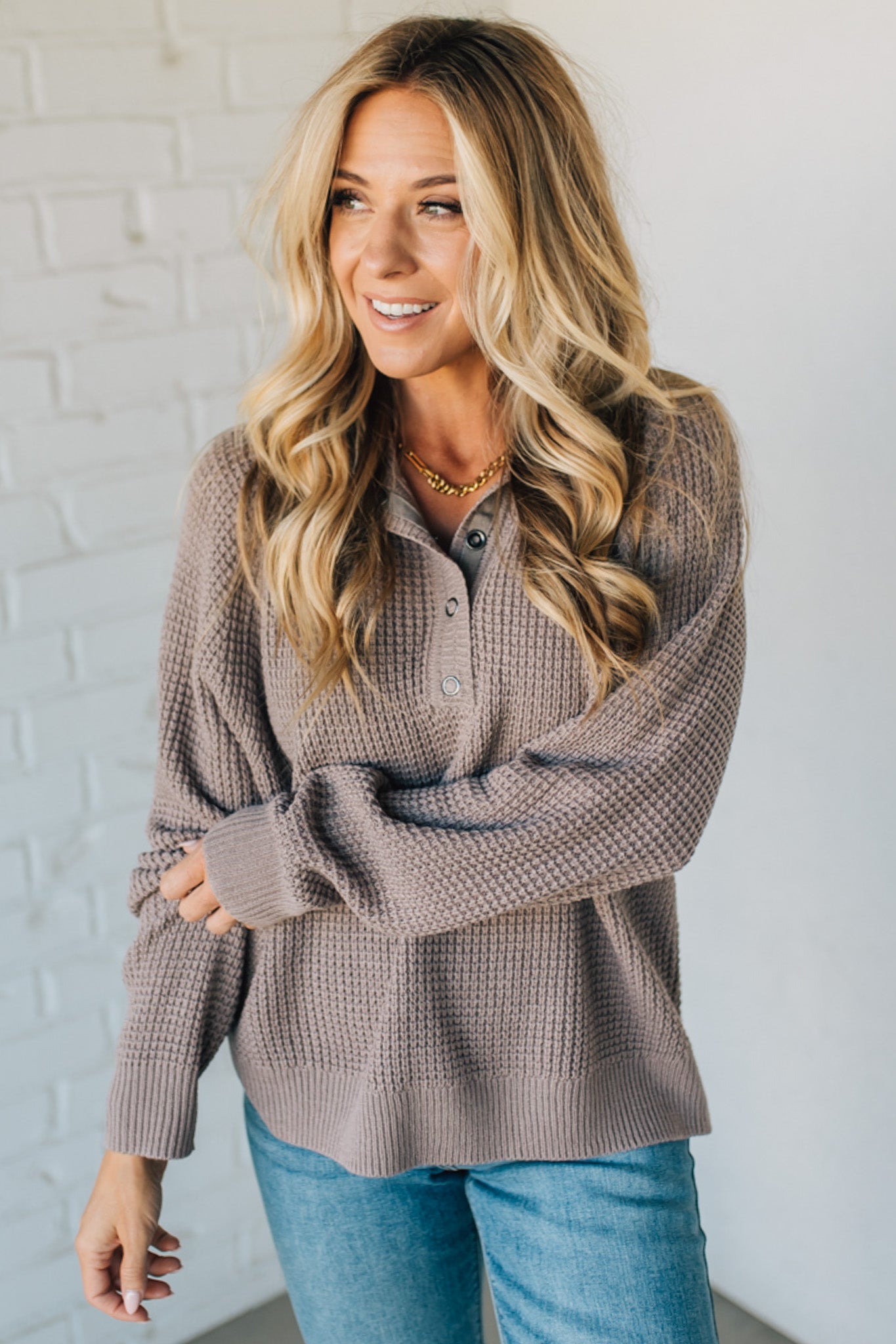 Woman wearing a brown textured sweater and blue jeans against a white brick wall.