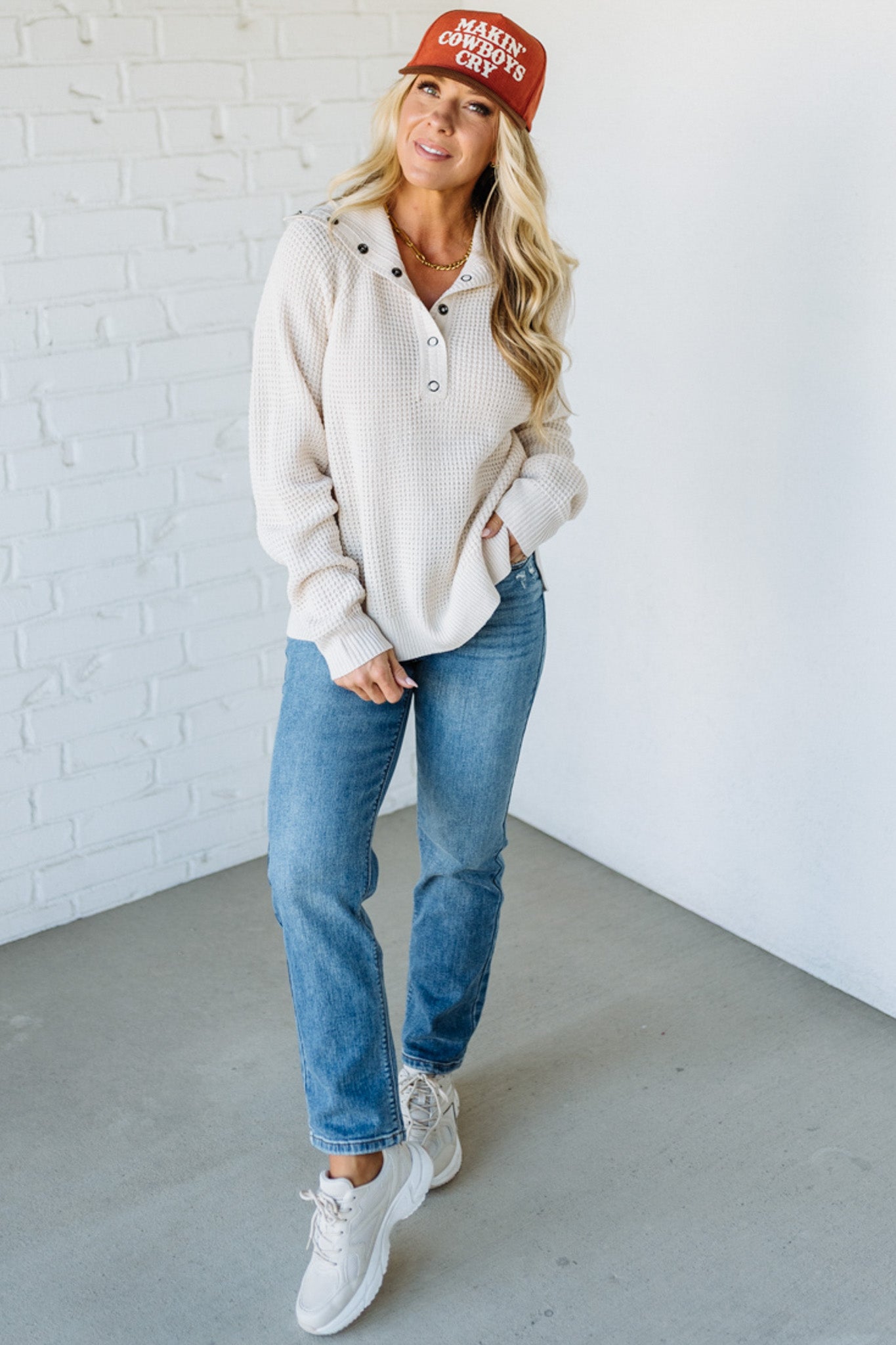 Woman wearing a beige sweater, blue jeans, and a red cap with text against a white brick wall.