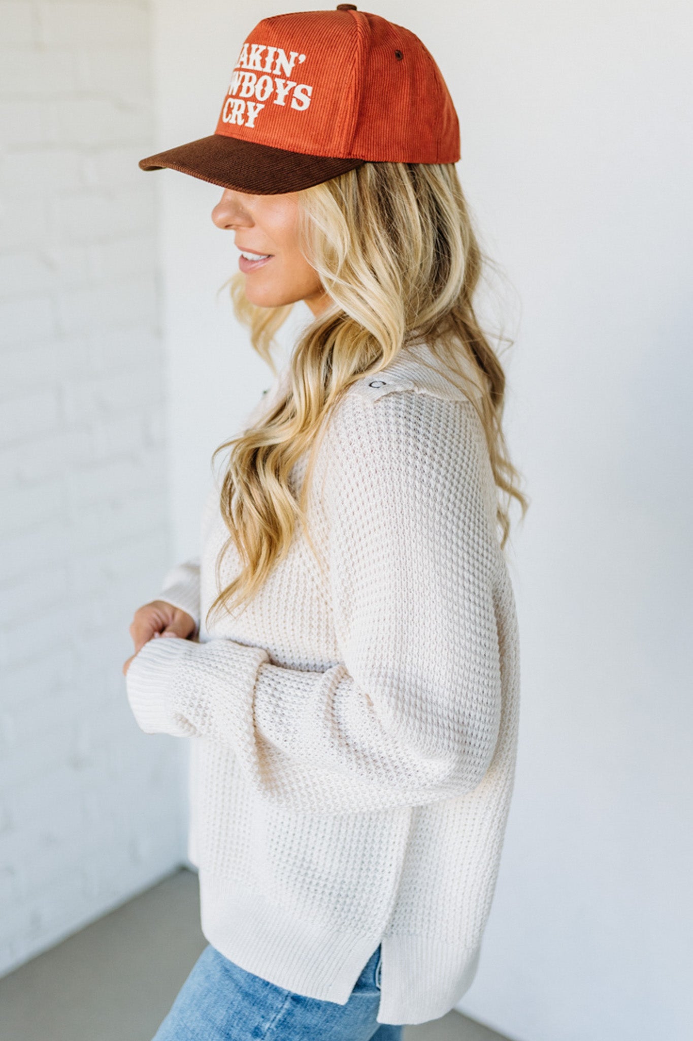 Woman wearing a red cap with text, white sweater, and blue jeans against a white background