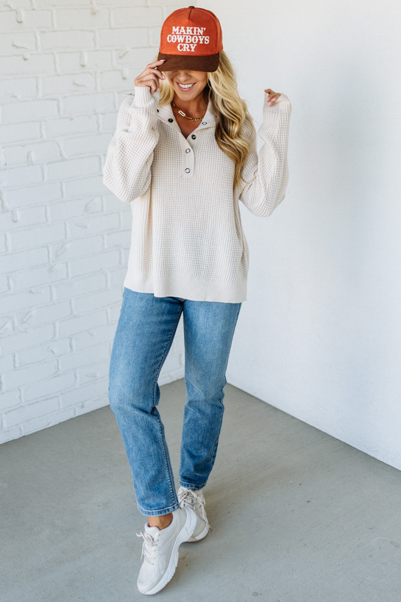 Woman wearing a white sweater, blue jeans, and a red cap with text, standing against a white brick wall.