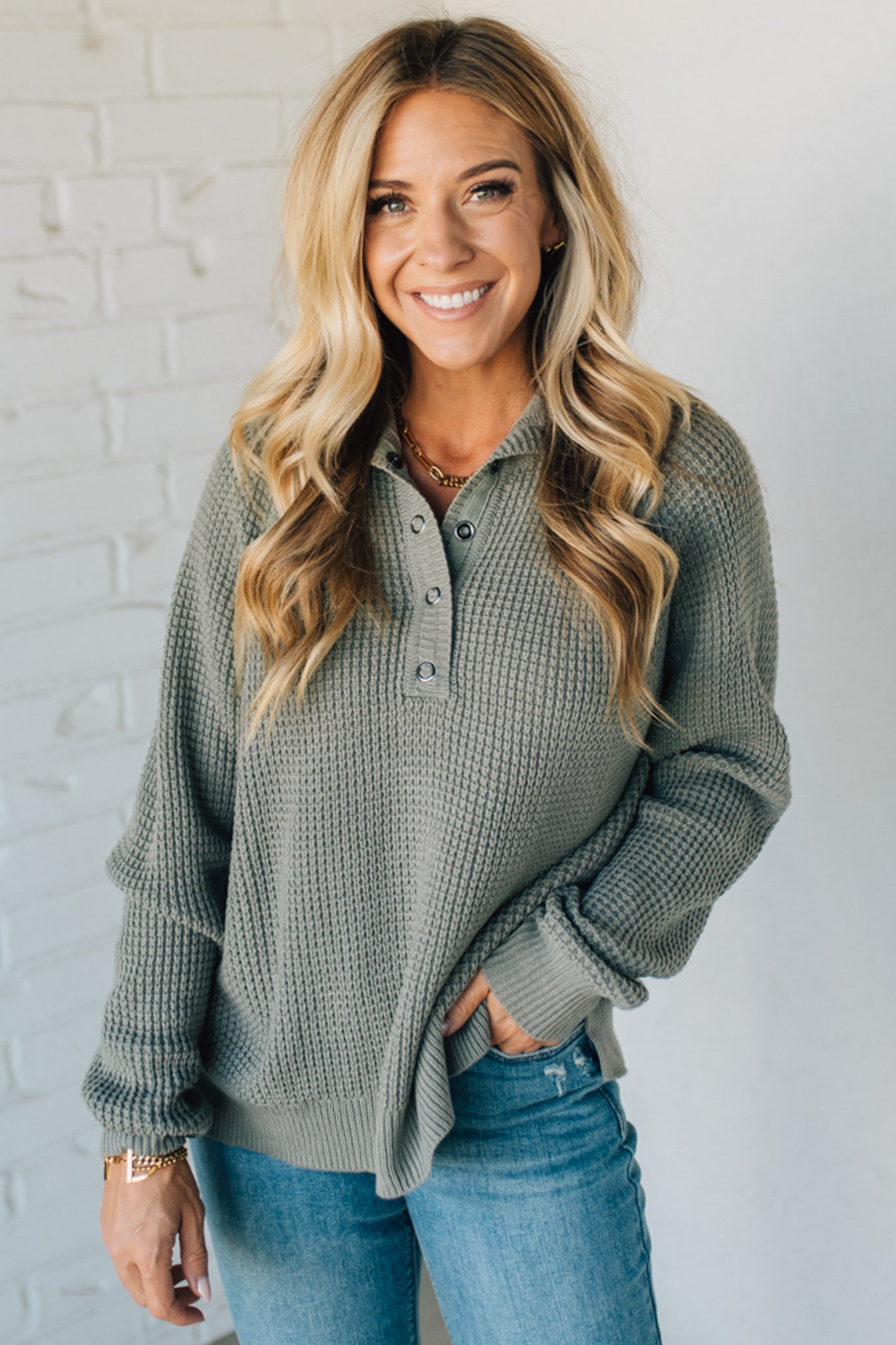 Woman wearing a green sweater and blue jeans against a white brick wall.
