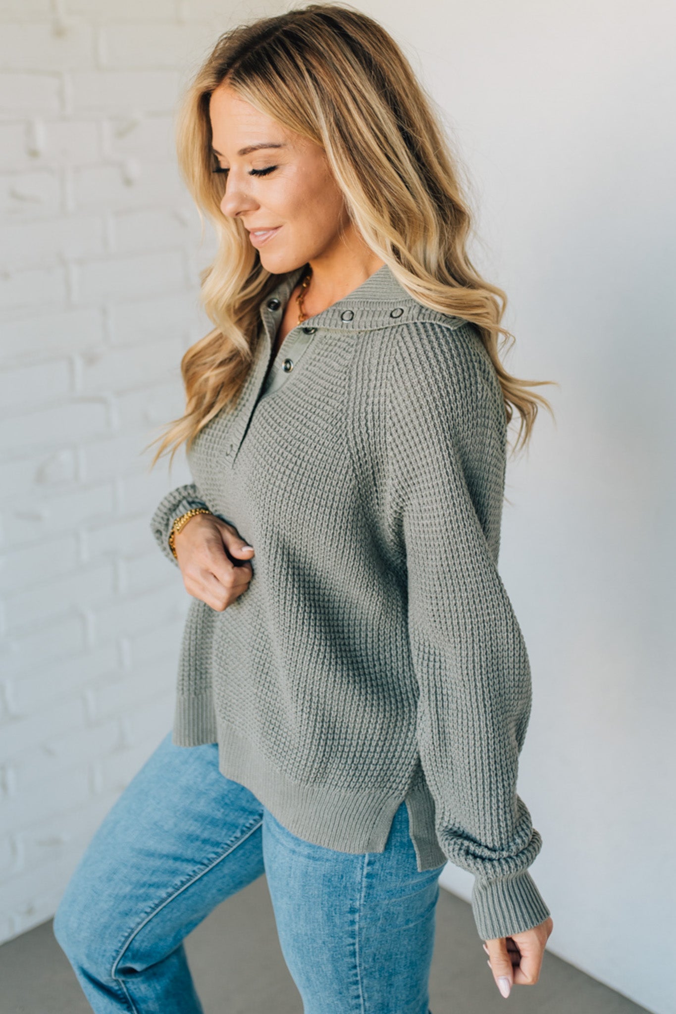 Woman wearing a green sweater and blue jeans against a white brick wall.