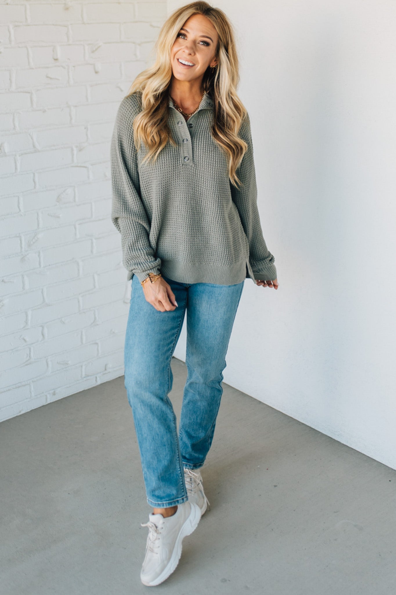 Woman wearing a green long-sleeve shirt and blue jeans standing against a white brick wall.