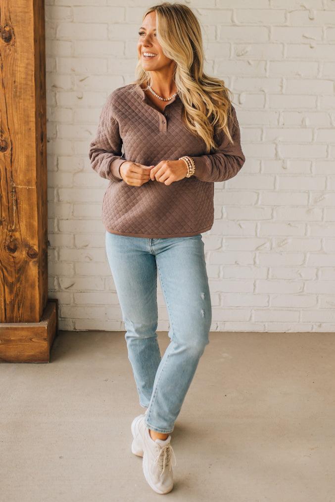 Woman wearing a brown sweater and light blue jeans standing against a white brick wall.